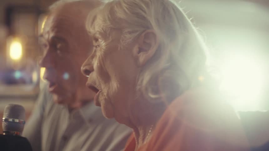 An elderly couple sits in their living room at midday with the woman in a wheelchair and the man beside her both singing into a microphone while laughing and smiling happily
