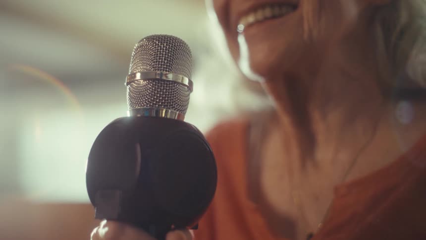 The first line shows a closeup of an elderly woman with white hair sitting in what appears to be a chair looking offcamera and talking with her mouth open she is wearing a rustcolored shirt and there is a professionallooking microphone in front of h