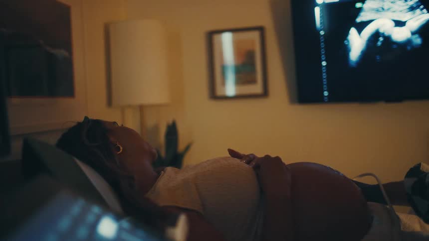 A pregnant woman lies on an examination bed with her hands resting on her belly watching the ultrasound images displayed on the monitor