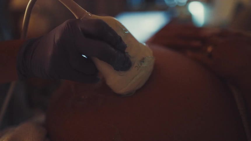 A pregnant woman is getting an ultrasound a person wearing a blue glove is holding the ultrasound probe and moving it across her belly which is covered in gel in the background an ultrasound machine with a brightly lit keyboard can be seen