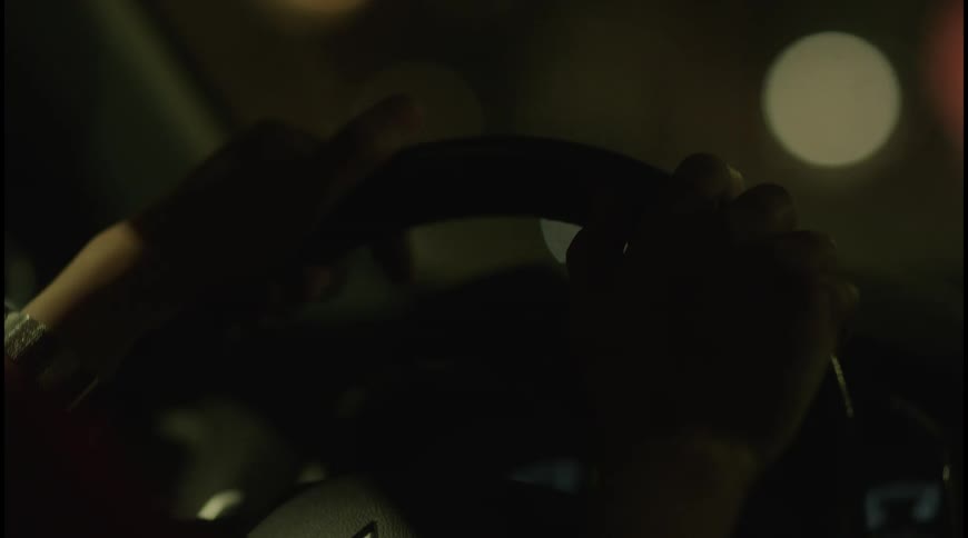 A drivers hands grip a steering wheel in the dark the background is a blur of outoffocus lights from the city at night
