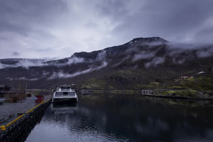 A small ferry is docked at a pier on a gray day with mistshrouded mountains towering in the background the still water reflects the overcast sky and the surrounding landscape