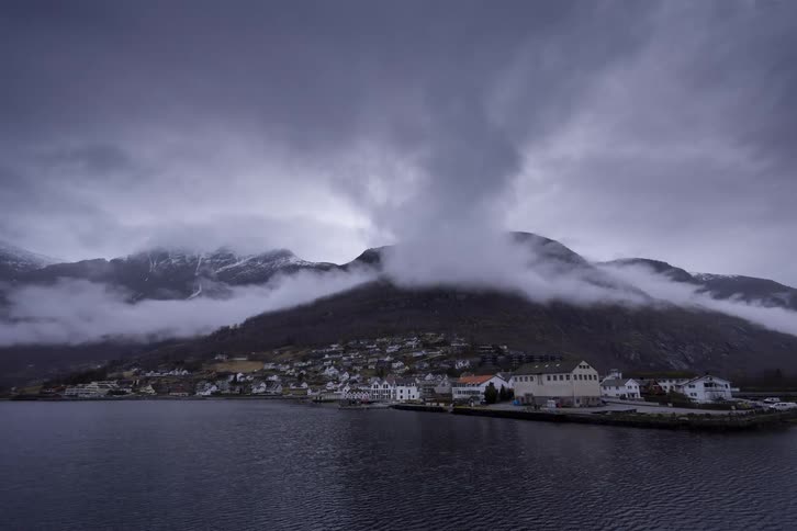 A small quaint village with white buildings sits at the base of a large misty mountain range a dark body of water is in the foreground with a pier jutting out from the shore