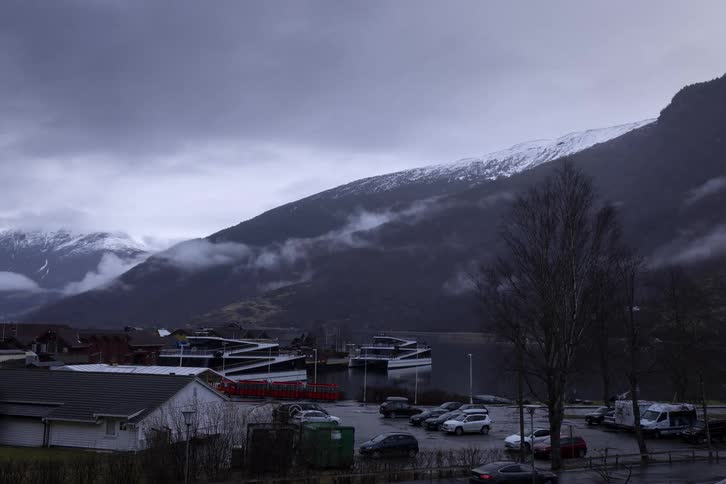 A landscape displays a mountain and a body of water with buildings and a parking area between them