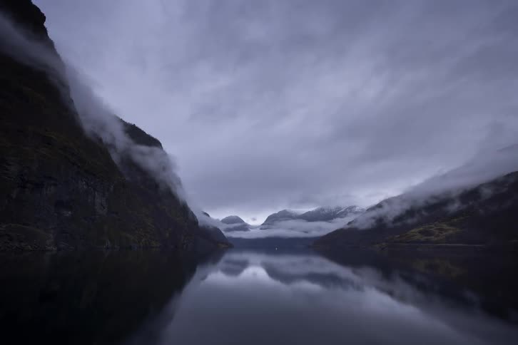A stunning wide shot captures a body of water in a fjord with dramatic mountains rising on either side the sky is filled with heavy dark clouds and a thick mist hangs over the mountains