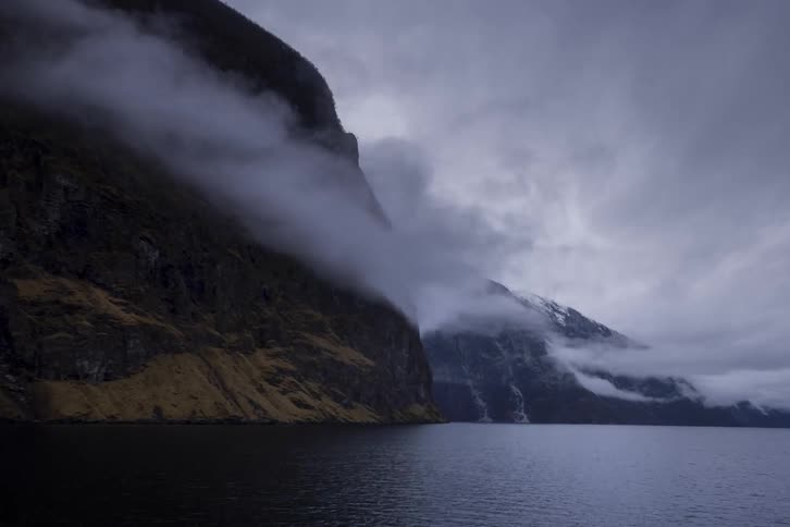 A boat moves slowly across a body of water in a fjord the scene is dominated by a sheer dark rock face on one side and a snowcapped misty mountain range on the other
