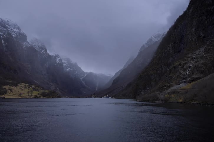 A drone shot captures a moody gray sky over a fjord with steep dark mountains on either side the drone slowly flies forward revealing a small village and several boats docked in the distance