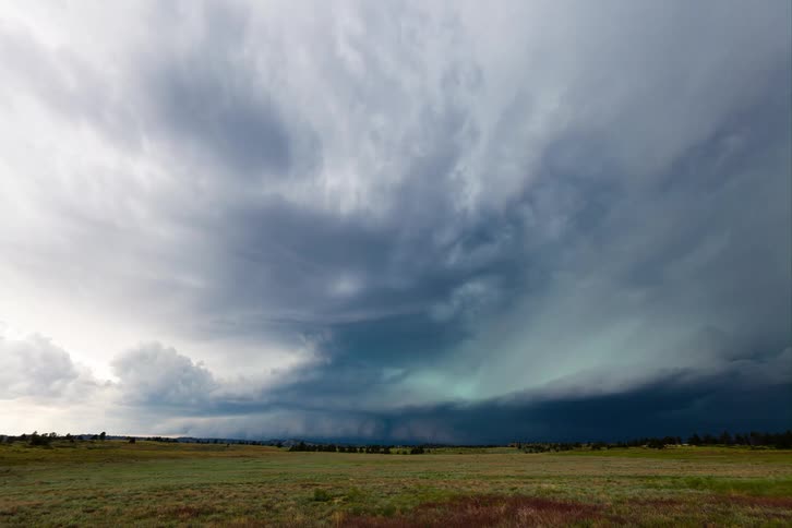 A vast dark storm cloud forms over a wide grassy field with a wall of rain falling in the distance the gray clouds fill the sky creating a dramatic ominous scene