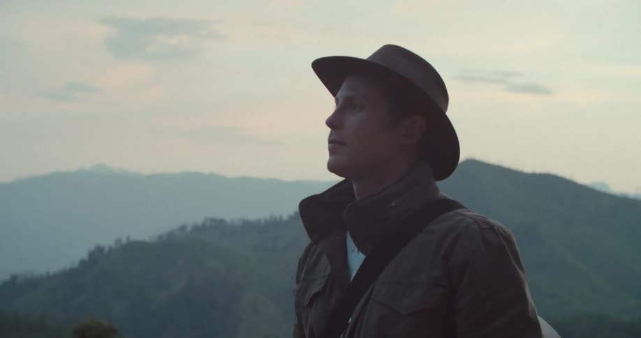 A silhouetted person wearing a cowboy hat and jacket stands with their back to the camera they look out at a vast mountainous landscape shrouded in mist