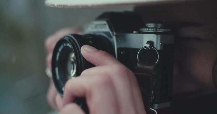A close view shows a persons hands holding a dark colored canon camera with their fingers turning the lens and pressing the button on top to take a photo