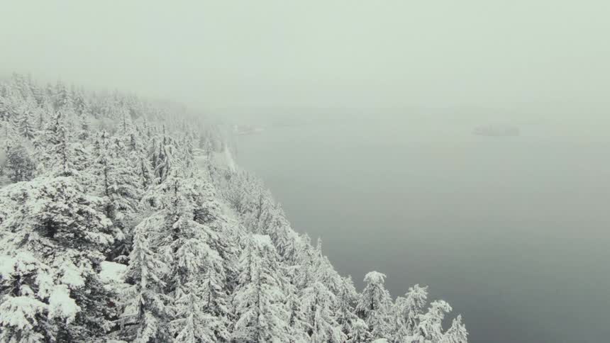 An aerial view of a snow covered forest shows a road on the left side and a large body of water on the right with a thick layer of fog covering the landscape and blurring the horizon