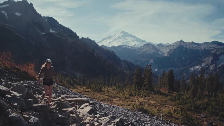 A hiker climbs a rocky trail below a snowy mountain peak
