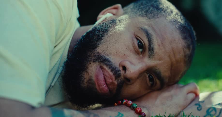 A bearded person with their eyes closed is lying down in the grass a bright beaded bracelet is visible on their wrist