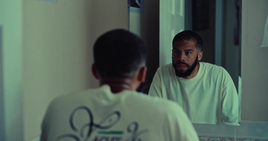 A person is seen from behind looking into a bathroom mirror their reflection stares back its expression shifting from a downward gaze to a direct serious look at the viewer