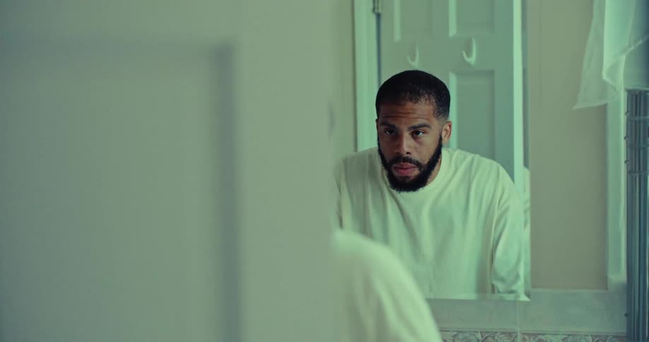 A man with a dark beard and a white shirt is seen in a mirror looking down and then raising his head to stare at his own reflection with a serious expression on his face