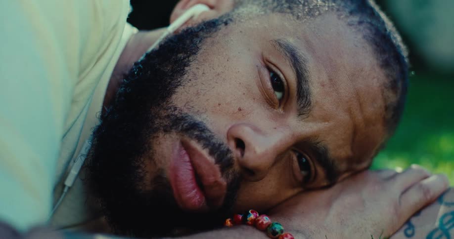 A person with a beard and a bright beaded bracelet rests their head on their arm in the grass the person is sleeping peacefully in the sunlight with their face in a slight profile