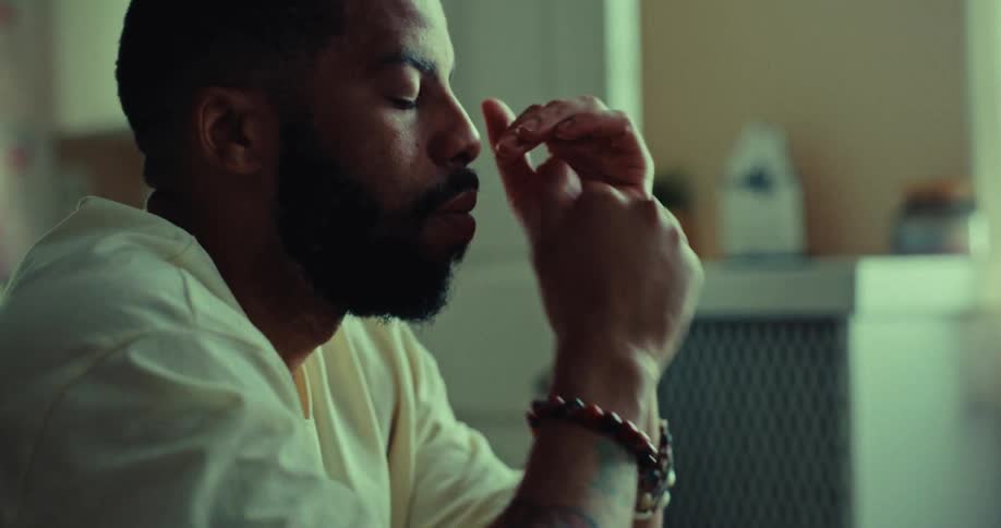 A bearded person is sitting with their head in their hand looking sad or worried they then bring their hands to their face looking distressed before placing them back on their lap