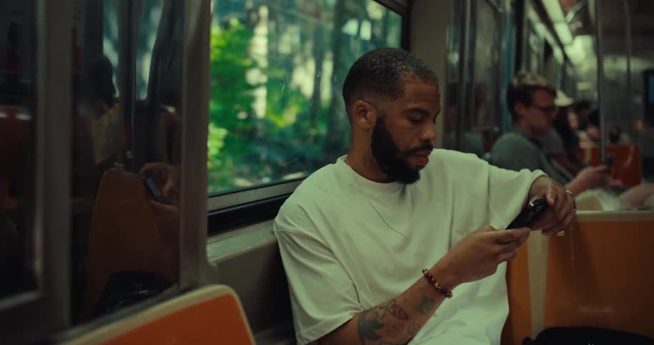 A man with a beard wearing a white shirt sits on a subway train holding a phone with sunlight and greenery visible through the window and other passengers in the background
