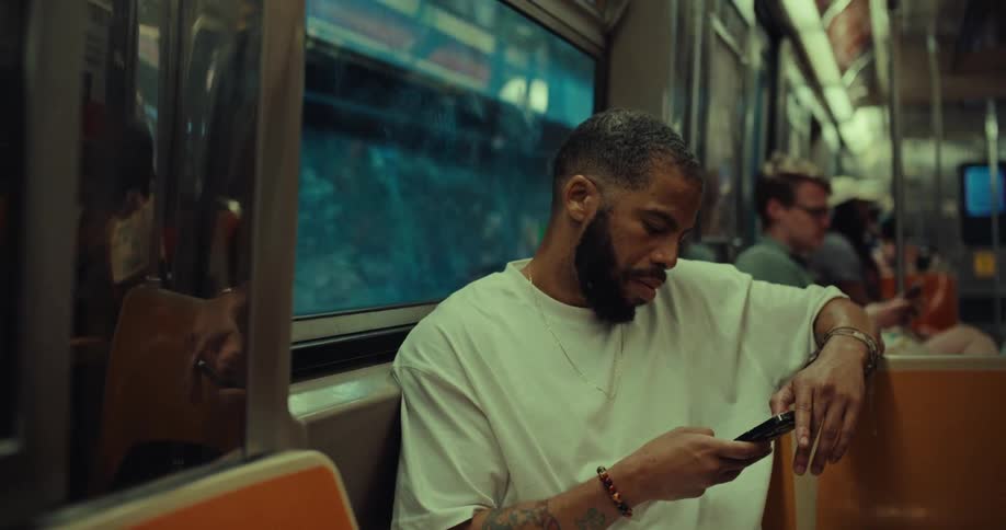 A bearded man with tattoos on his arms is riding a subway car sitting down and looking at his phone he appears to be texting or scrolling his face illuminated by the screen