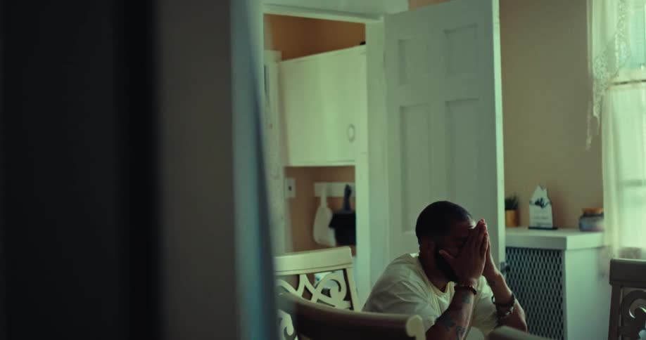 A man with a short beard and a white shirt is sitting at a wooden table in a dimly lit room with his head in his hands as he looks down at the table with a sad expression on his face