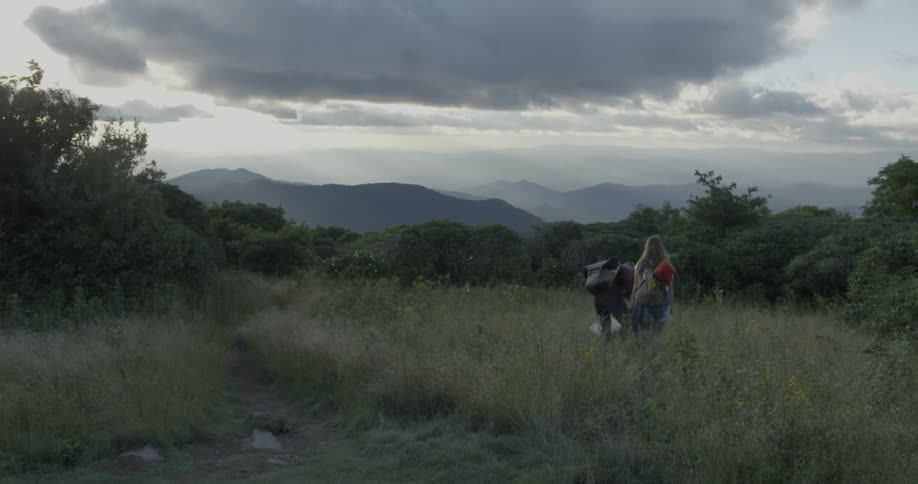 Two people walk in the tall grass with camping equipment