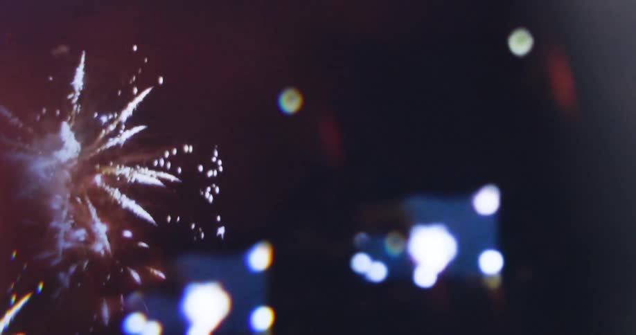 A shot from the side shows a person holding up a phone and taking a video there are blurry bright lights and what appear to be fireworks in the background