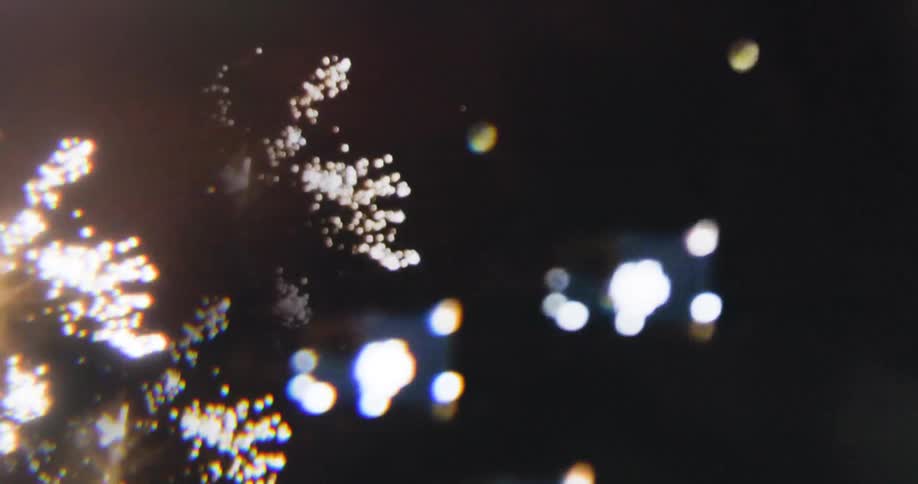 A man wearing glasses uses his phone to take pictures of fireworks bursting in the sky and illuminating the environment at night
