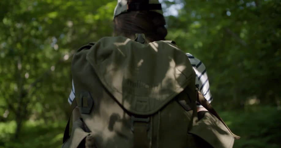 A hiker wearing a striped shirt cap and green backpack walks along a bright forest trail