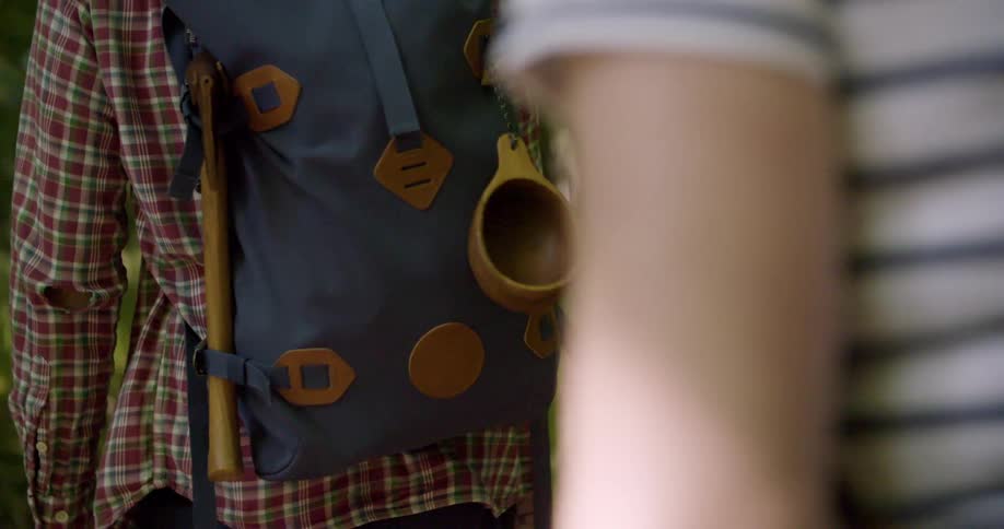 A close view from the side shows a man hiking in a forest with a backpack an axe and a wooden cup attached to it