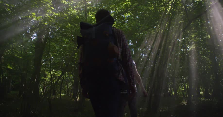 A man and a woman with backpacks are hiking through a dense forest with bright sun rays filtering through the tall trees
