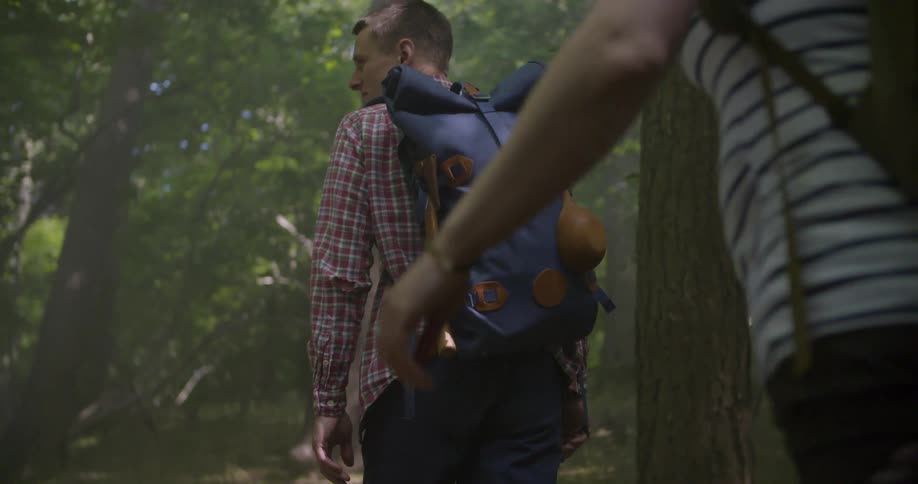 A person with a blue backpack walks through a shaded forest trail partially framed by another figure in the foreground