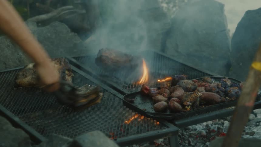 A closeup shot shows a person using tongs to flip a large piece of meat on a grill over hot coals beside it smaller pieces of meat and vegetables are also cooking with smoke rising from the sizzling food