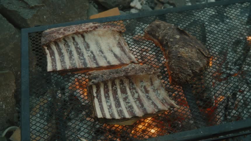 Large cuts of meat including racks of ribs cook over an open flame on a metal grill