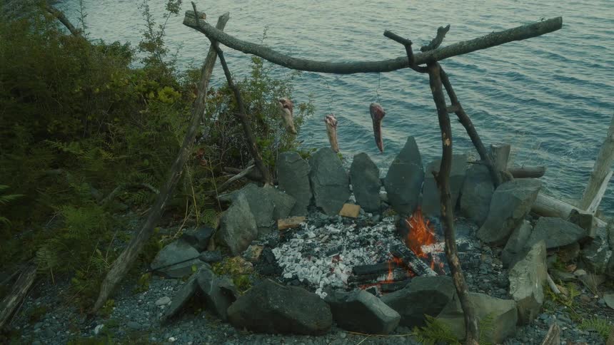A campfire burns by the riverbank with meat suspended on a wooden stick roasting over the flames at midday