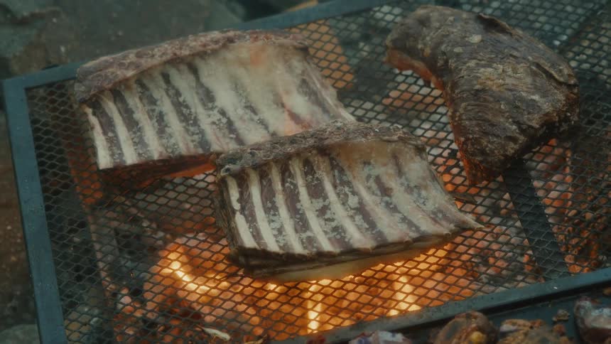 Two racks of ribs and a large cut of meat grill over open flames on a metal grate
