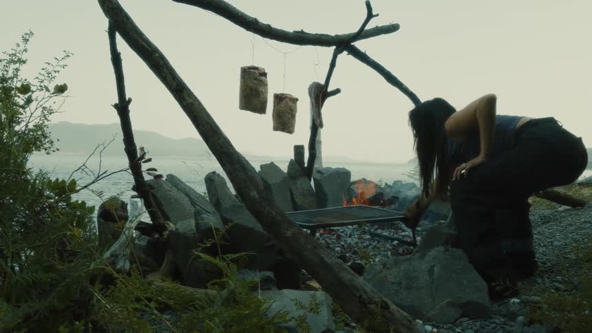 A woman is placing a grill rack over a rocky campfire on a beach with a view of the ocean and mountains