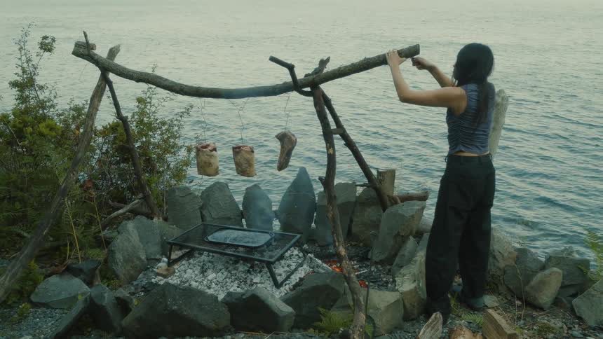 Woman secures wire to long piece of wood with three pieces of meat hanging from it over a fire