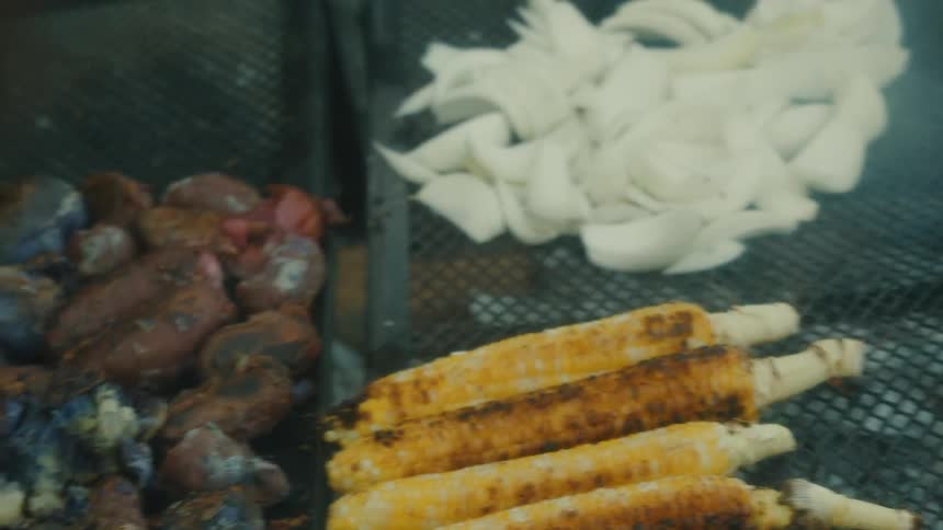 Pieces of meat being roasted with vegetables on the grill over an outdoor fire pit