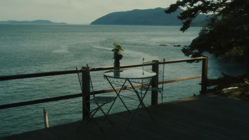 A beautiful view of a tranquil sea framed by distant misty mountains and a wooden deck with a small table and chairsthe scene captures a serene moment on a seaside deck where a gentle breeze rustles the leaves on a small plant overlooking the vast