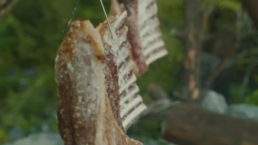 The first shot is a closeup of a large piece of meat being cooked over an open fire it appears to be a whole fish or a very large cut of meat salted and hung from a hook the shot slowly pans down to show more of the meat which is brown and textured