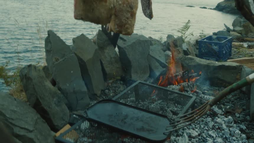 Pieces of meat suspended with coarse salt being roasted over a fire by a lake