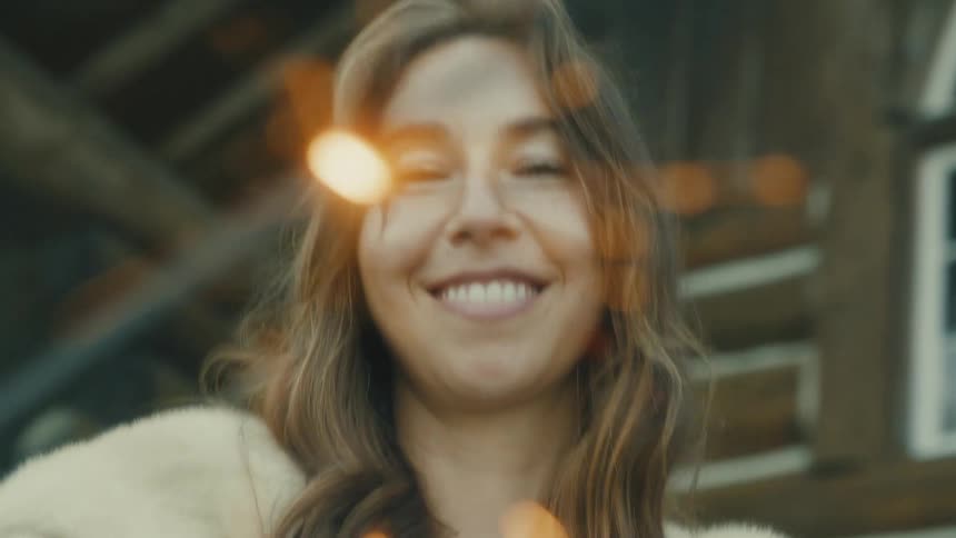 A smiling young woman with long brown hair and a white fluffy sweater is laughing happily while standing outdoors in the sunlight
