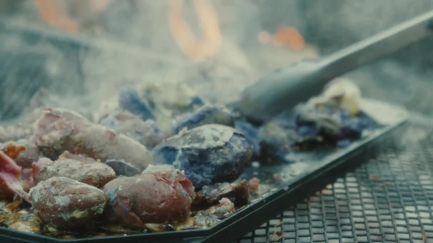 A hand with tongs turns pieces of meat and potatoes on a grill over a fire