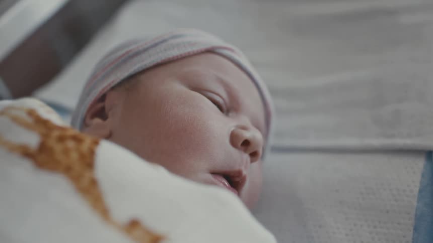 A newborn baby wrapped in a blanket and wearing a hat lies on the bed at midday