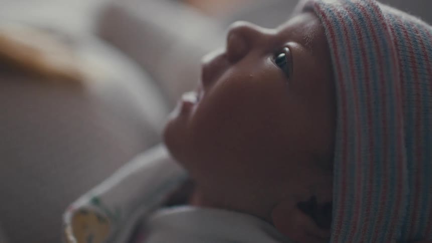 A mother holds her baby indoors at midday the child wearing a small hat and gazing around with a curious expression as soft daylight filters through the room