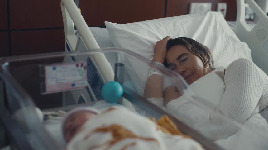 A tired but happy young woman is lying in a hospital bed looking at her newborn baby sleeping in a clear plastic crib next to her