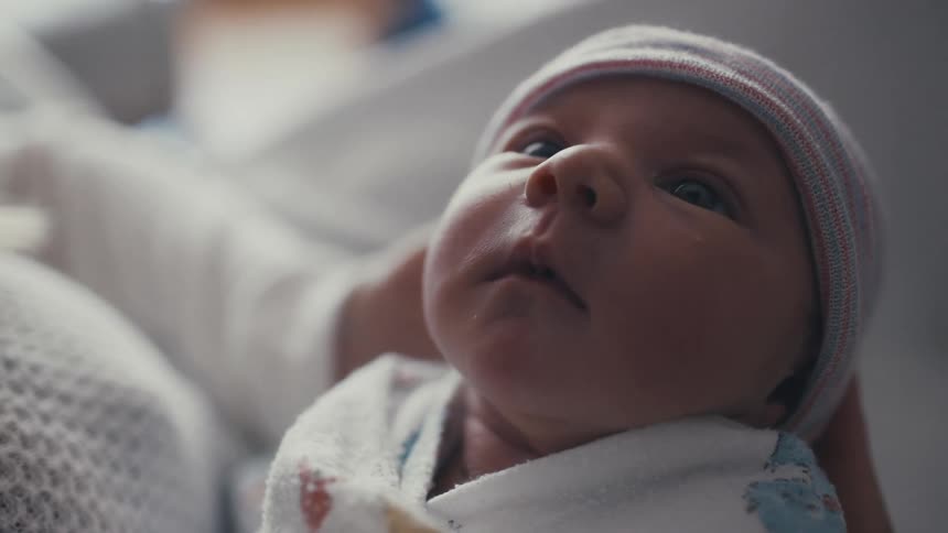 A close view of a newborn baby with wide blue eyes wearing a knit beanie and wrapped in a blanket with its mouth open