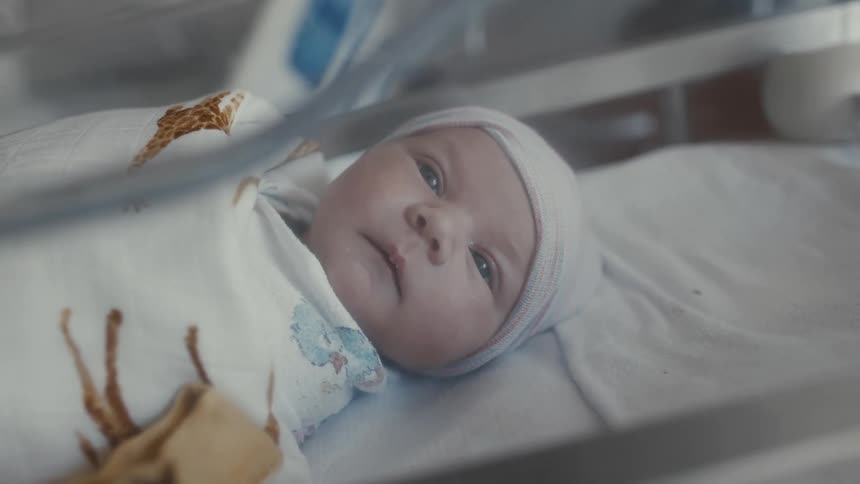 A close view of a newborn baby with a knit beanie and a blanket with a giraffe pattern lying in a hospital crib and looking directly at the camera
