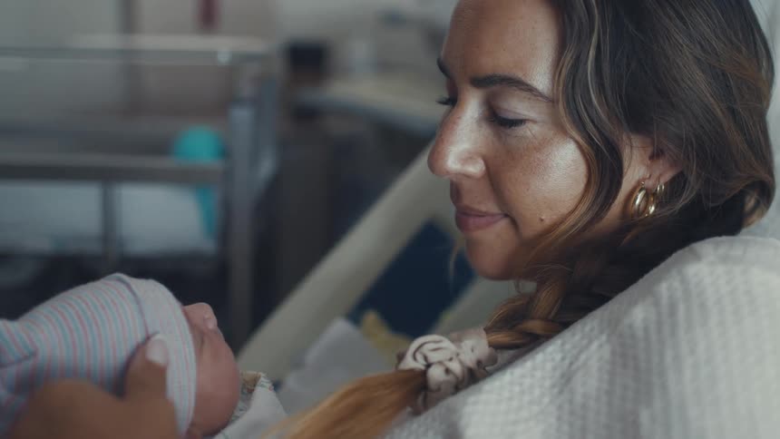 A woman with a braid cradles her newborn in a hospital bed gazing at the baby with tenderness