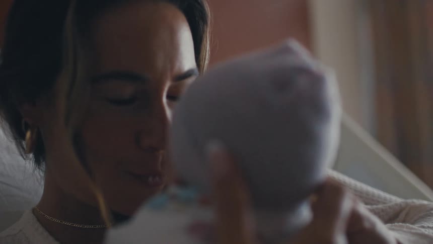 A young woman is lying on the bed holding her infant and smiling as she playfully teases the baby